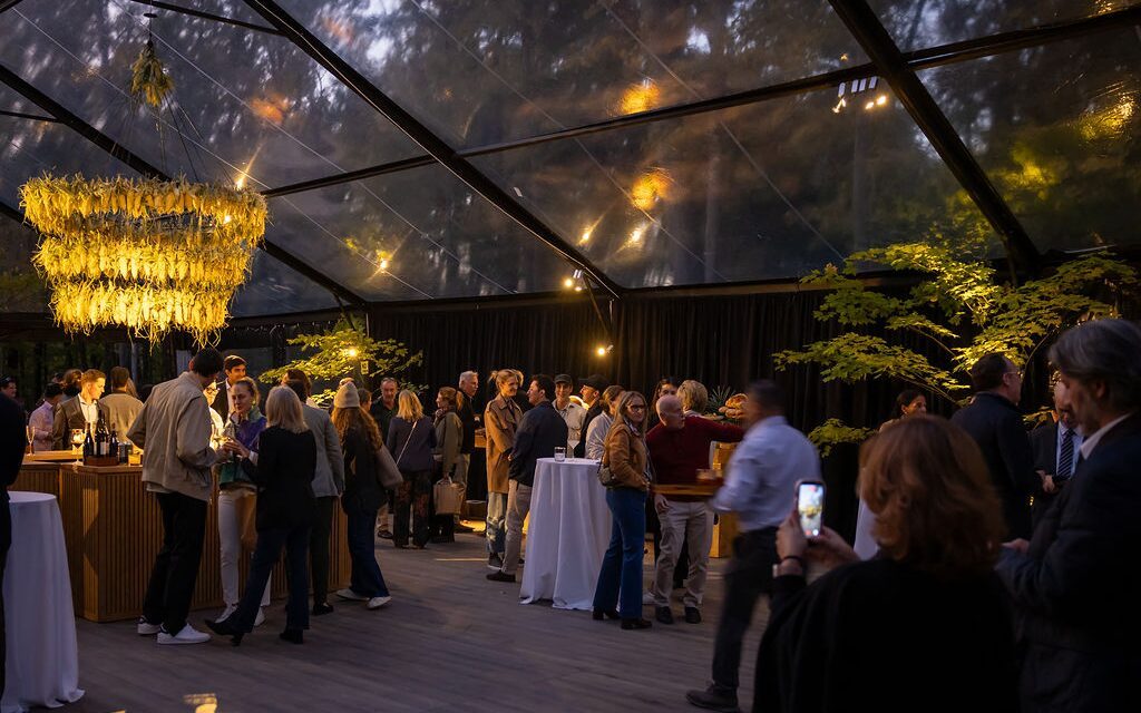 Guests in the clear pavilion beneath the tree canopy at dusk, the corn-husk chandelier glowing overhead